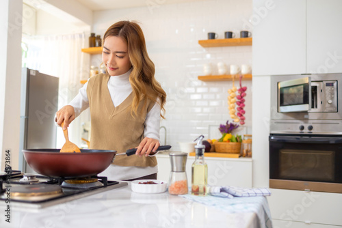 Young beautiful Asian woman enjoy cooking healthy food and pasta in cooking pan on stove in the kitchen at home. Happy female having dinner meeting party celebration with friends on holiday vacation.