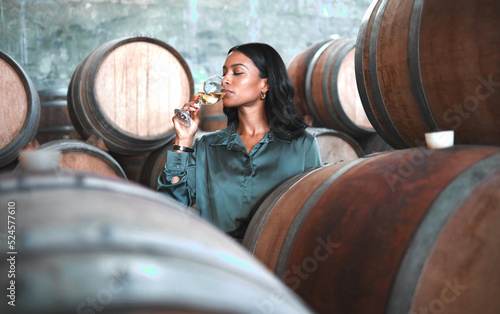 Woman doing wine tasting, drinking glass of chardonnay or sauvignon blanc in winery cellar amongst barrels on vineyard. Beautiful oenologist or sommelier enjoying a relaxing, luxury beverage indoors.