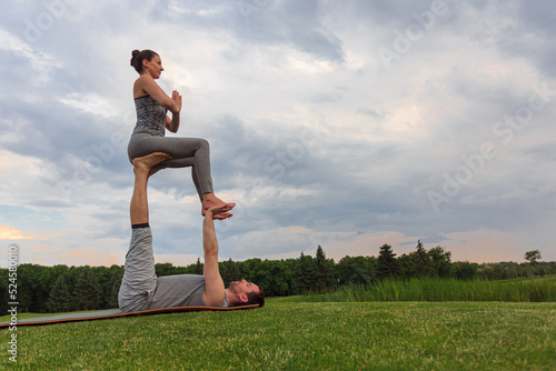 Healthy man lying on grass and balancing woman. Couple doing acrobatic yoga exercise in park