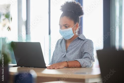 Covid, face mask and a black woman working on a laptop in a modern office. Sick businesswoman on the internet at work. College student, online exam and in campus library, protected against the virus.
