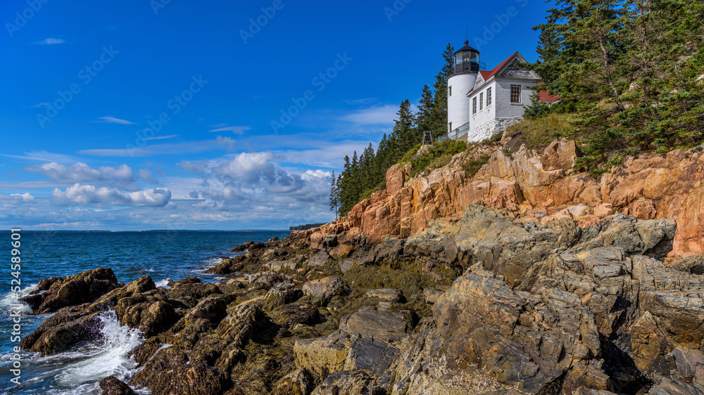 Bass Harbor Head Light - A wide-angle view of Bass Harbor Head Lighthouse standing on top of colorful seaside cliff against blue sky on a sunny Autumn morning. Acadia National Park, Maine, USA.