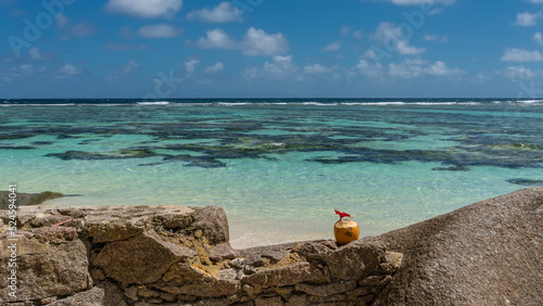 On a granite boulder there is a yellow coconut decorated with a red tropical flower. Ahead - turquoise ocean, blue sky with clouds. Seychelles. La Digue Island