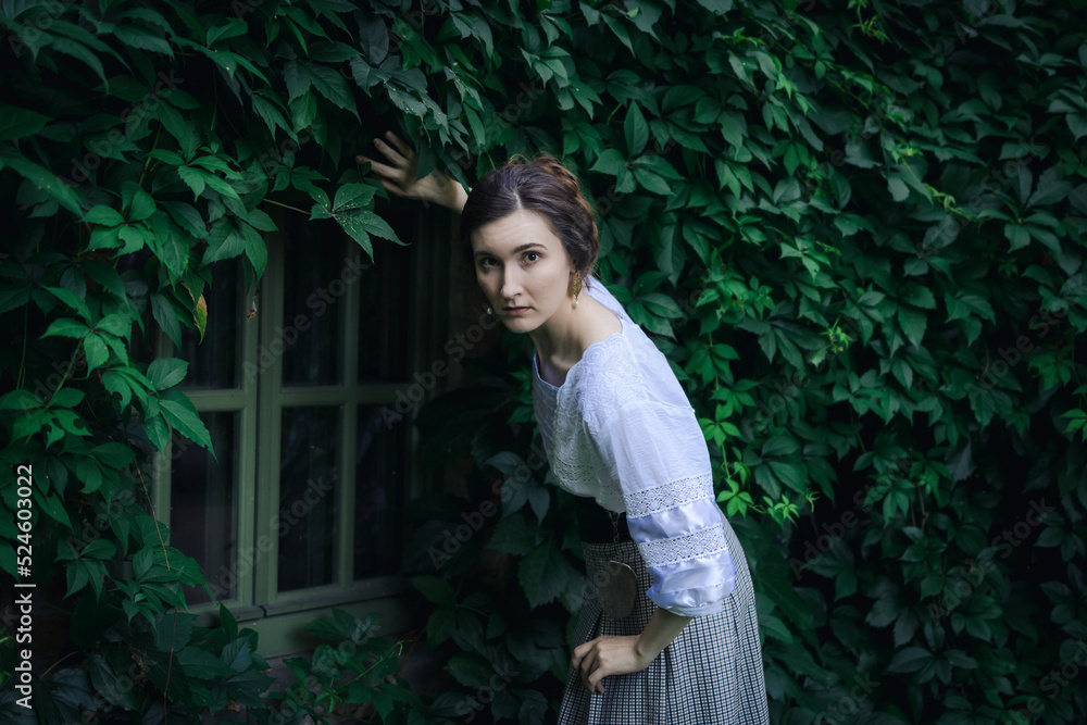 Portrait of a young slender woman in a 1910s costume. The lady peers through the ivy-covered window. Portrait of a young slender woman in a 1910s costume. The lady peers through the ivy-covered window.