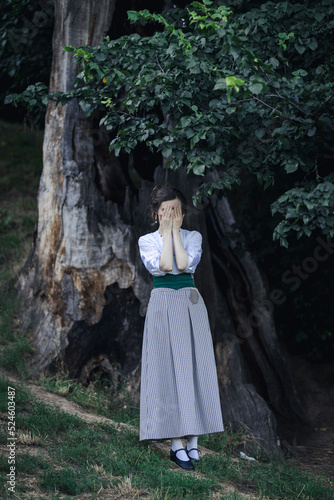 Full-lenght portrait of a young slender woman in a 1910s costume. Lady stands under an old broken tree, covering her face with her hands.