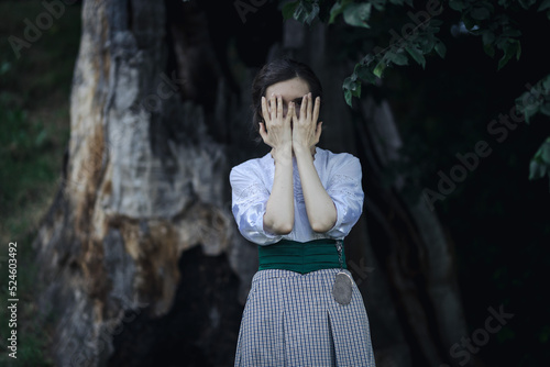 Portrait of a young slender woman in a 1910s costume. Lady stands under an old broken tree, covering her face with her hands photo