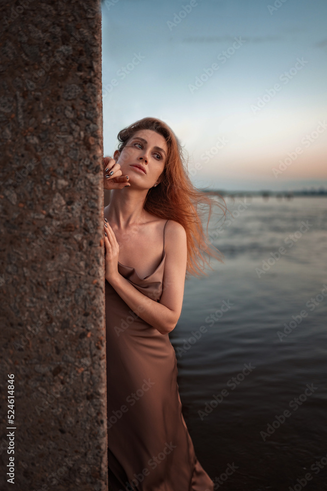 Naklejka premium A beautiful slender girl with red hair and a perfect figure stands in the river at sunrise in a pink and gold satin tight-fitting sundress with lowered straps. Gentle sexuality, romance and femininity