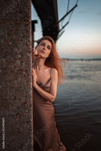 A beautiful slender girl with red hair and a perfect figure stands in the river at sunrise in a pink and gold satin tight-fitting sundress with lowered straps. Gentle sexuality, romance and femininity