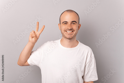 Hipster cheerfully smiling at the camera while he is showing the peace sign standing near a grey wall wearing a white t-shirt