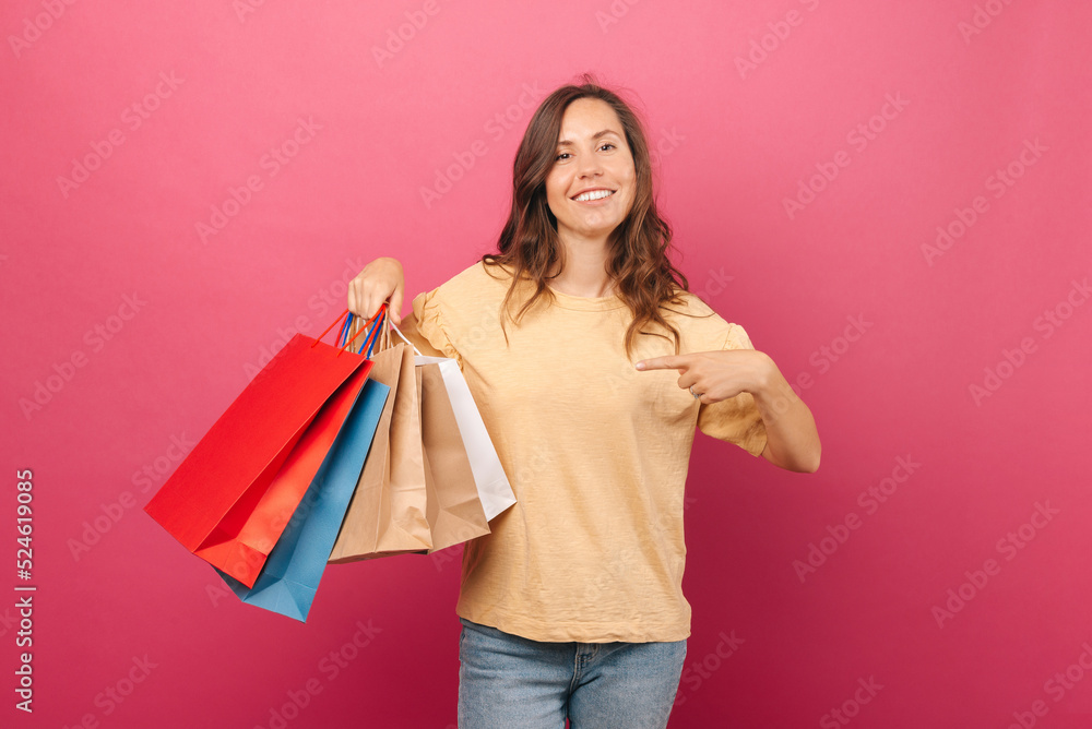 Young female is smiling at the camera and holding some shopping bags. She is being pleased with her shopping, wears a nude shirt near a pink background