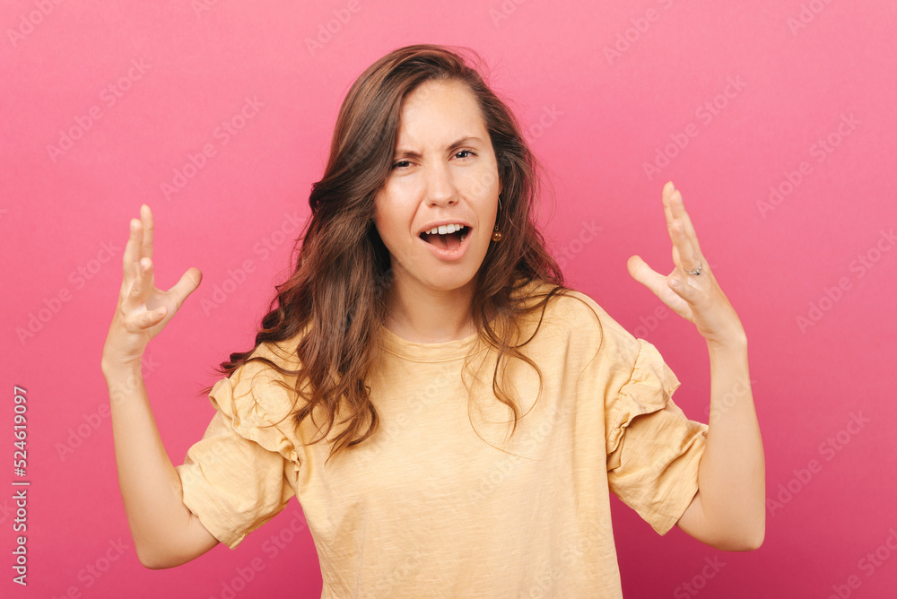 Confused woman with brown hair is looking directly at the camera with both her hands up wearing a nude shirt near a pink background Confused woman with brown hair is looking directly at the camera with both her hands up wearing a nude shirt near a pink background