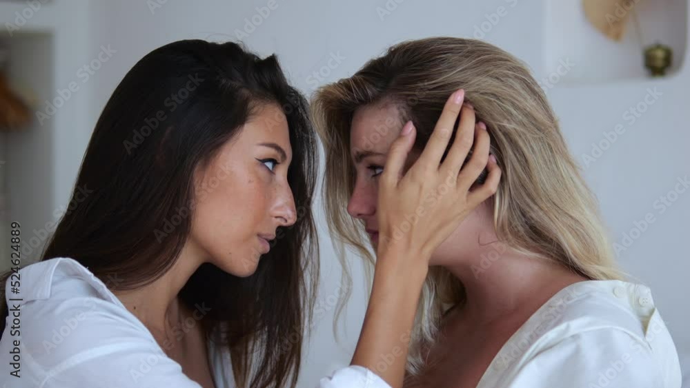 Two sensual lesbians are sitting on the bed in front of each other, the brunette runs her hand into the blonde's hair and gently touches her face. The concept of love in same-sex marriage of lesbians. Two sensual lesbians are sitting on the bed in front of each other, the brunette runs her hand into the blonde's hair and gently touches her face. The concept of love in same-sex marriage of lesbians.