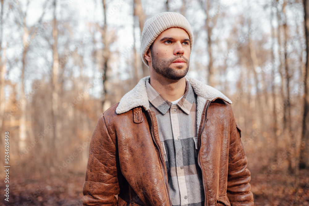 A portrait of a young white man posing in the woods dressed in autumn clothes: brown jacket, checkered canadian style shirt, off-white beanie, looking to the right A portrait of a young white man posing in the woods dressed in autumn clothes: brown jacket, checkered canadian style shirt, off-white beanie, looking to the right