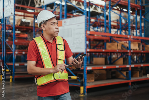 Warehouse Workers standing and holding tablet in Logistic center. Asian Male Manager wearing safety vest and Hard Hat to working about shipment in storehouse, Working in Storage Distribution Center.