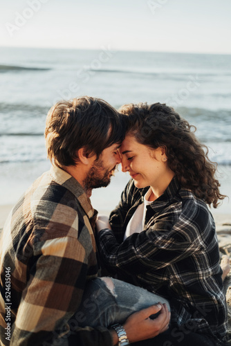 Close up of young woman and man hugging while sitting together on the seashore with sea waves on the background at sunrise