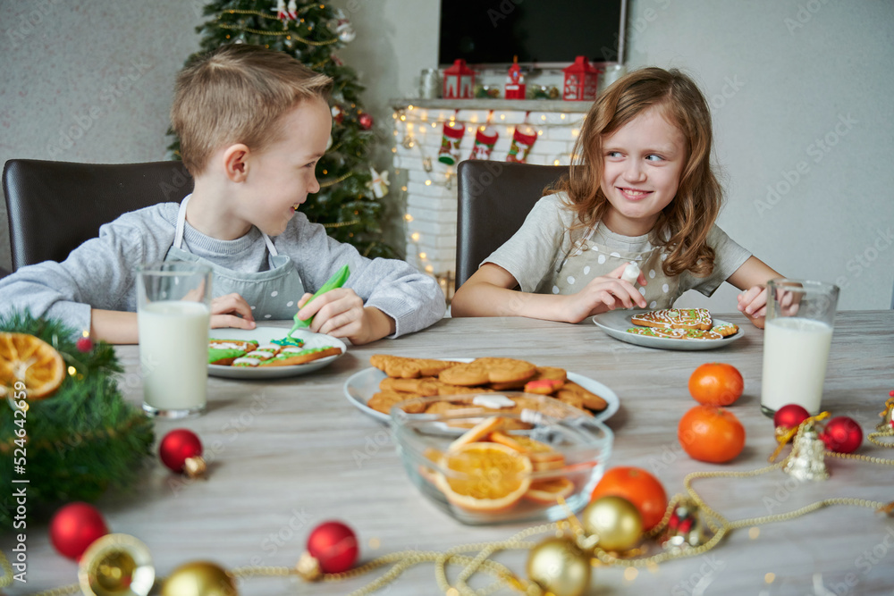 Sibling children decorate honey gingerbread cookies on table in living rom. Icing of Christmas baking. Concept Christmas holiday. Two glasses of milk on the table for Santa.