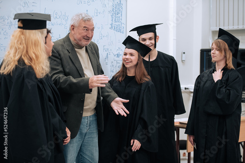 In the math class, an elderly gray-haired professor congratulates students in robes and caps on the completion of their studies and hugs them