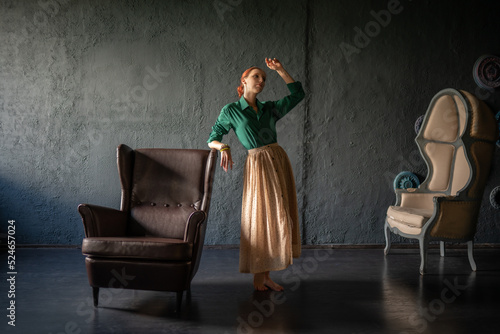 Beautiful red-haired girl in a green shirt and a long linen skirt, in a vintage interior, stands next to an old armchair. An Italian farmer in an Italian-style villa. Natural beauty and tendernes