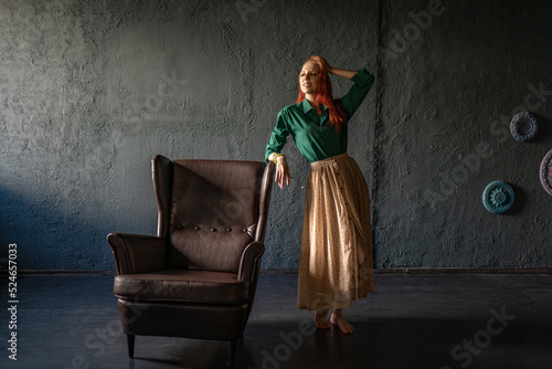 Beautiful red-haired girl in a green shirt and a long linen skirt, in a vintage interior, stands next to an old armchair. An Italian farmer in an Italian-style villa. Natural beauty and tendernes photo