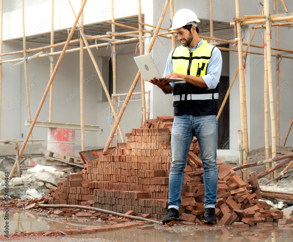 Bearded man engineer wears safety helmet inspecting housing development project at construction site using laptop computer. Contractor manager examining residential building estate infrastructure.