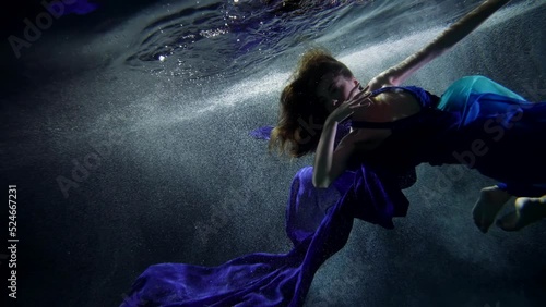 a woman in a blue dress with a long train under the surface of dark water with bubbles. the camera is tilted