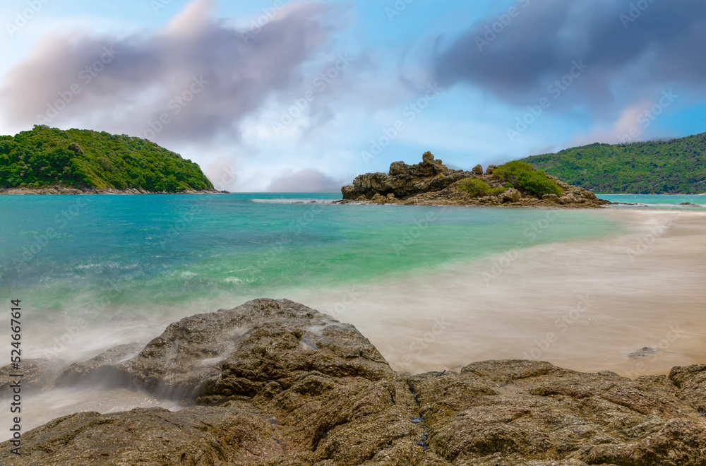 Fototapeta premium Long exposure of waves crashing on the rocks with beautiful island and mountains in Background. photographed in Naiharn Nai Harn Beach Phuket Thailand