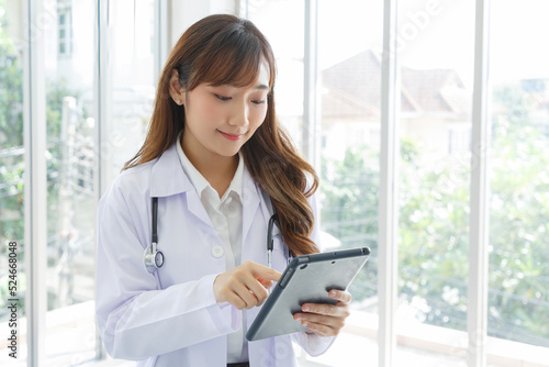 Beautiful and cheerful good mood young lady asian doctor dressed uniform smiling arm stethoscope while using tablet on lobby hospital background.