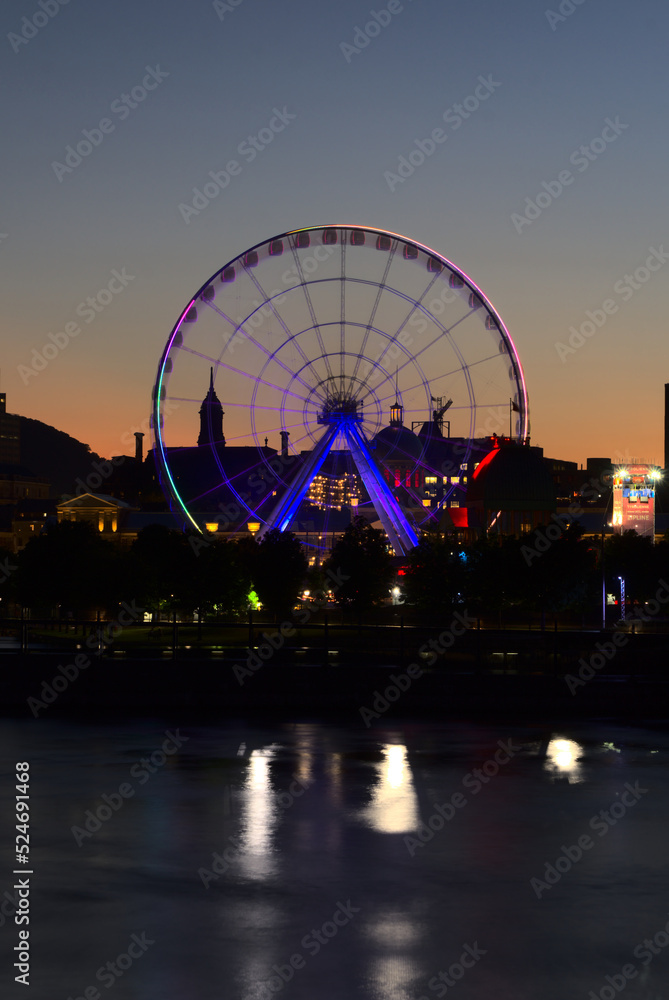 Long exposure of Montreal Grand Ferris Wheel in Old Port with motion blurat twilight blue hour. Reflections of sun rays on Saint Laurent river with a background of cloudy blue sky