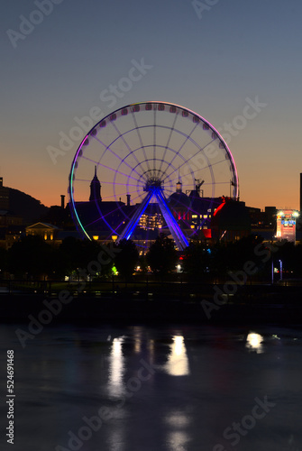 Long exposure of Montreal Grand Ferris Wheel in Old Port with motion blurat twilight blue hour. Reflections of sun rays on Saint Laurent river with a background of cloudy blue sky