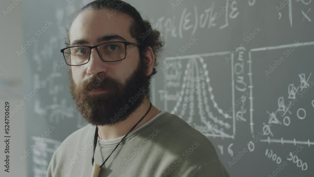 Portrait chest up shot of bearded biracial student in eyeglasses posing for camera against chalkboard in university classroom Portrait chest up shot of bearded biracial student in eyeglasses posing for camera against chalkboard in university classroom