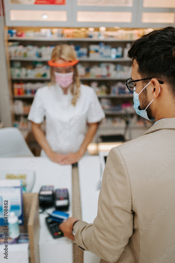 Young business man choosing and buying drugs in a drugstore while talking with attractive female pharmacist. She helping him with expert advice. They are wearing protective face masks.