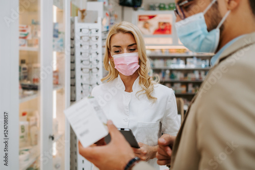 Young business man choosing and buying drugs in a drugstore while talking with attractive female pharmacist. She helping him with expert advice. They are wearing protective face masks.