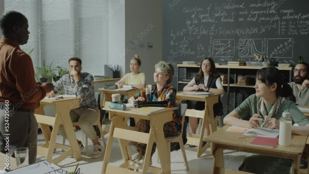 Group of multiethnic college students sitting at desks in classroom and listening to African American professor during lecture Group of multiethnic college students sitting at desks in classroom and listening to African American professor during lecture