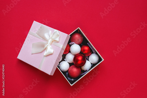Top view on round Christmas decorations in pink open gift box on red background. Beautiful red and white round toys. Concept of Christmas and New Year or romantic Valentines Day. Copy space for text.