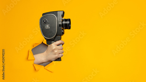 Hand holding small video old retro black camera 8 mm in paper torn hole in yellow background with copy space.