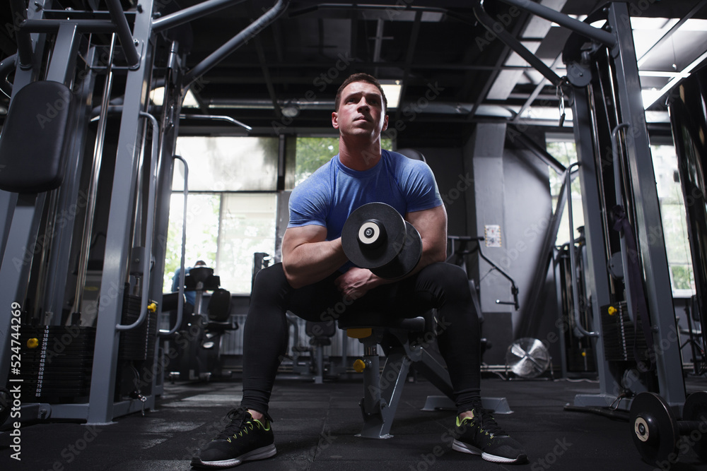 Full length shot of a muscular sportsman on weight training at gym, doing biceps curls with dumbbell Full length shot of a muscular sportsman on weight training at gym, doing biceps curls with dumbbell