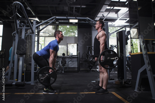 Profile full length shot of twin brothers bodybuilder doing barbell deadlift workout, standing face to face at gym