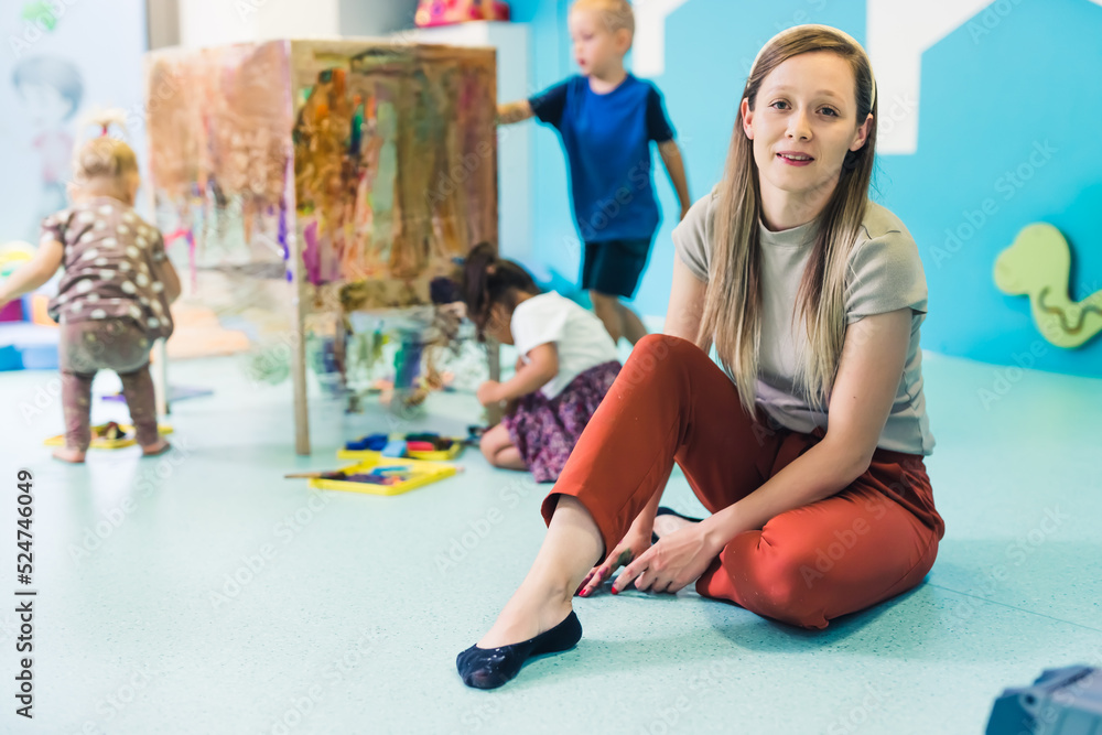 Naklejka premium Young caucasian day-care-center teacher sitting on the floor and looking at the camera while on the background toddlers painting with tempera paints on a cling film wrapped around the wooden shelving
