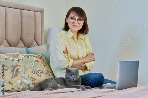 Portrait of middle aged woman with cat sitting on bed at home with laptop