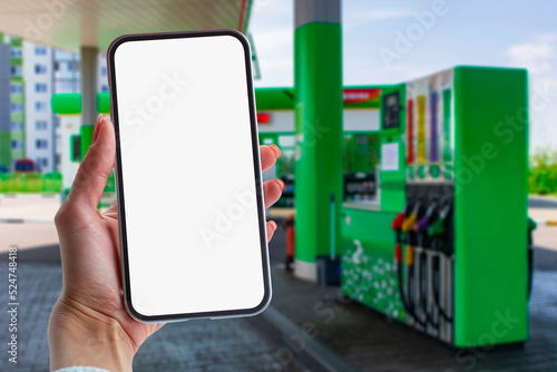Woman holds a close-up of a smartphone with a white screen in his hands against the backdrop on a gas station. Technology mockup for apps and websites.