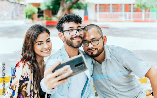 Three smiling friends sitting on a bench taking a selfie. Front view of three cheerful friends taking a selfie while sitting on a bench. Three friends taking a selfie on a park bench, Friendship conce