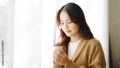 Young asian woman standing beside window and holding mug in living room at home, She drinking milk after wake up in the morning © Monster Ztudio