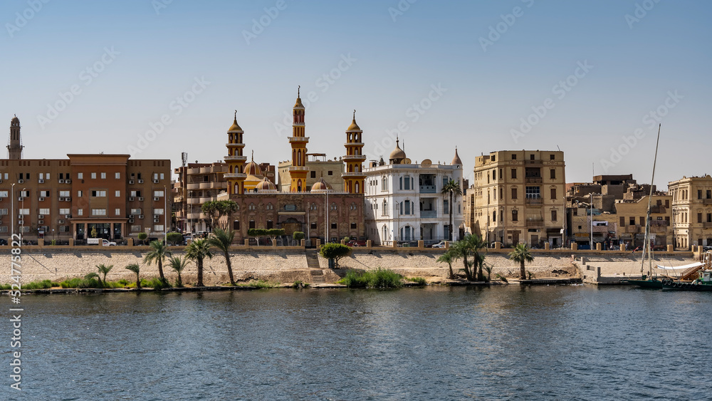 The embankment of the Nile in Egypt. City buildings against a clear blue sky. Palm trees grow at the water's edge. A stone staircase descends to the river