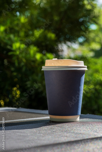 Coffee break with a take away paper cup full of hot coffee placed on a grey plastic placemat. Sunlight in the morning shines with a nature view in a background. Relax, cozy and warm feeling.