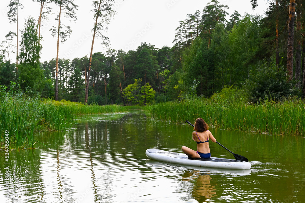 A woman drives on the Sup Board through a narrow canal surrounded by dense grass. Active weekend vacations wild nature outdoor. A woman is sitting cross-legged.