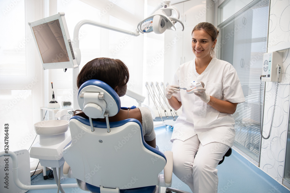Fototapeta premium Smiling dentist teaching young female patient to clean teeth with visual aid and toothbrush. African American woman having consultation in dental clinic. Oral hygiene concept