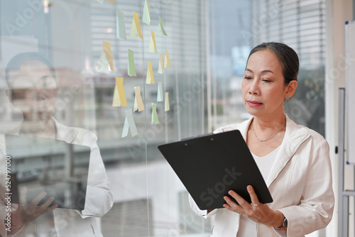 Foto Asian senior old lady businesswoman looking on chart board and standing beside the window, There is a view of tall buildings in the big city in the background