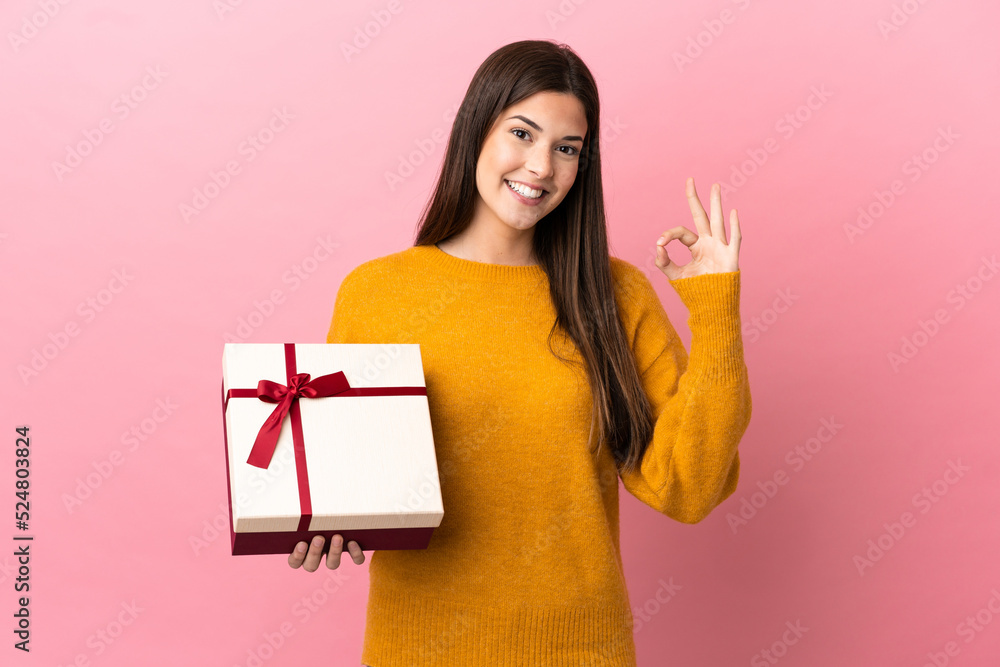 Teenager Brazilian girl holding a gift over isolated pink background showing ok sign with fingers Teenager Brazilian girl holding a gift over isolated pink background showing ok sign with fingers