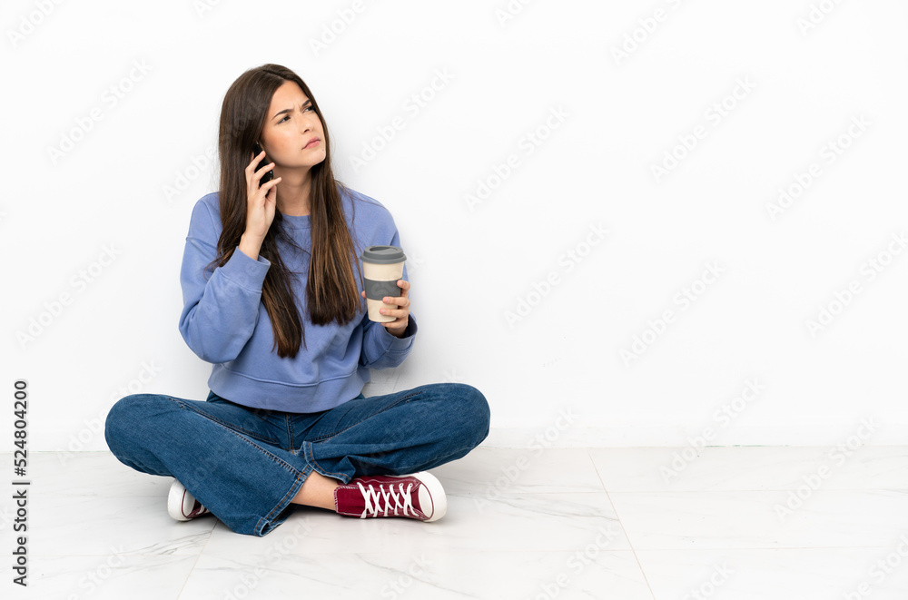 Young woman sitting on the floor holding coffee to take away and a mobile Young woman sitting on the floor holding coffee to take away and a mobile