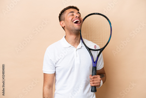 Young brazilian handsome man playing tennis isolated on beige background laughing © luismolinero