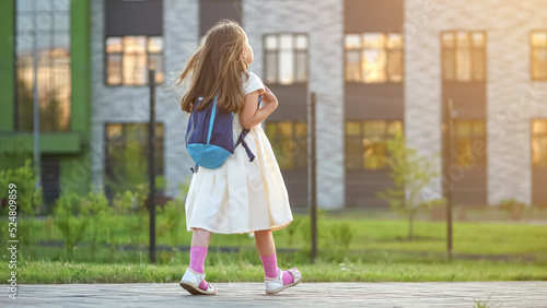 Slika na platnu Preschooler girl with blue backpack walks past big school building